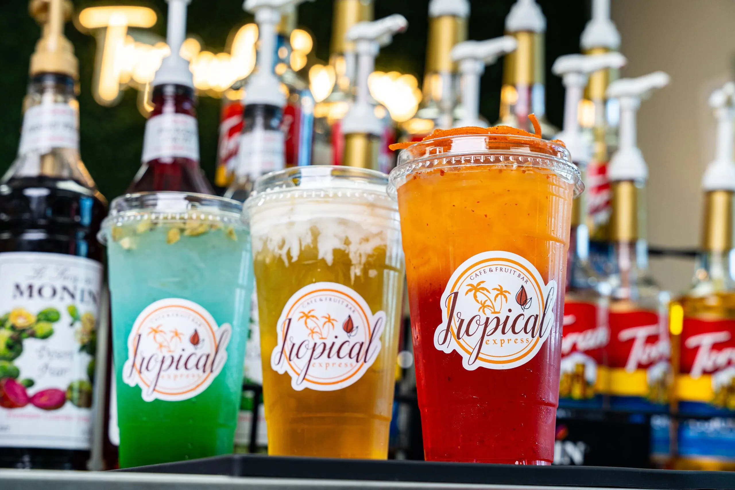 A lineup of vivid green, gold, and red Tropical Express iced drinks on the bar in front of rows of flavor syrups