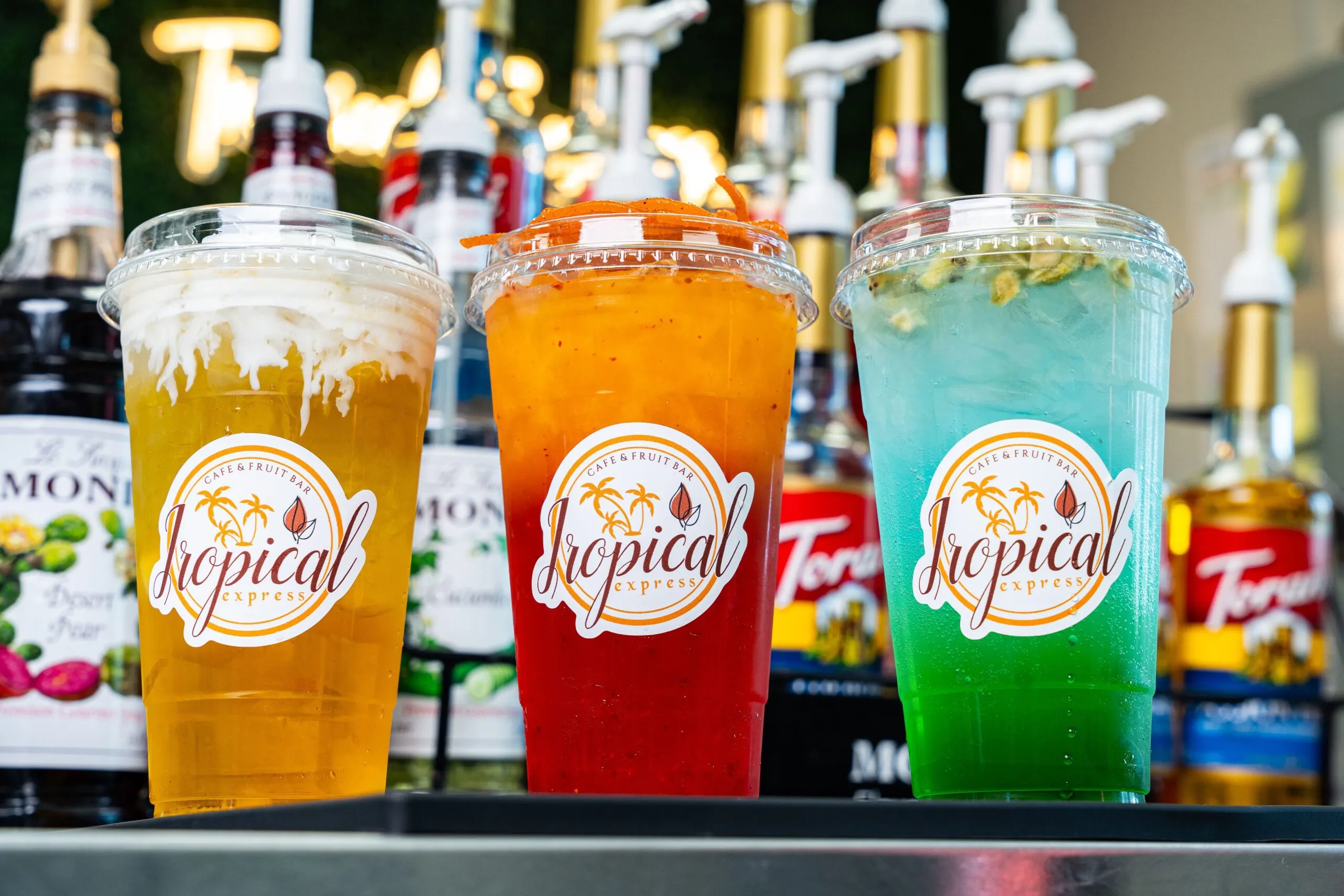 Three Tropical Express branded cups of colorful iced drinks — amber, red, and blue-green — lined up in front of flavor syrup bottles
