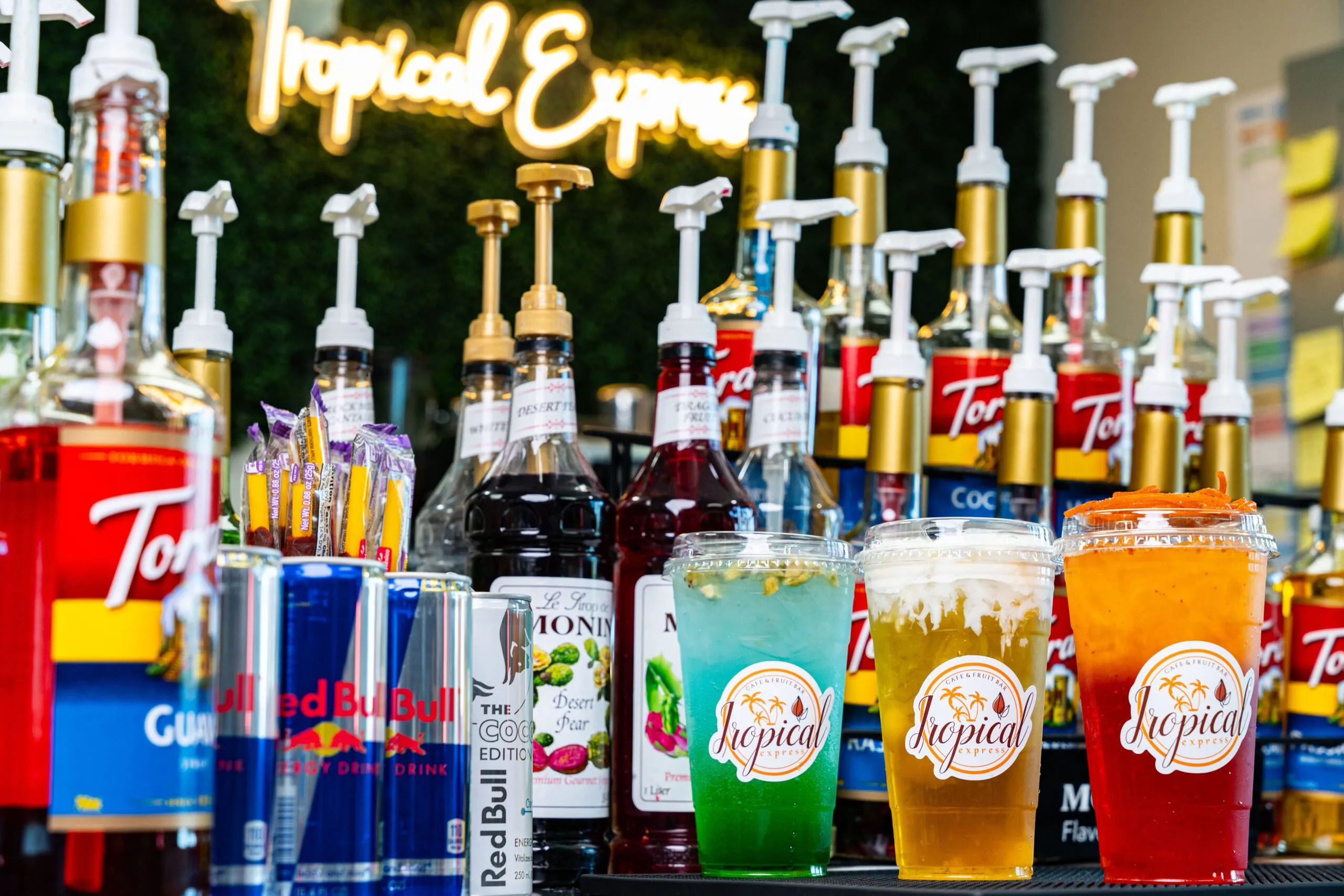 Tropical Express energy drinks beside Red Bull cans and flavor syrups, under the glowing Tropical Express neon sign