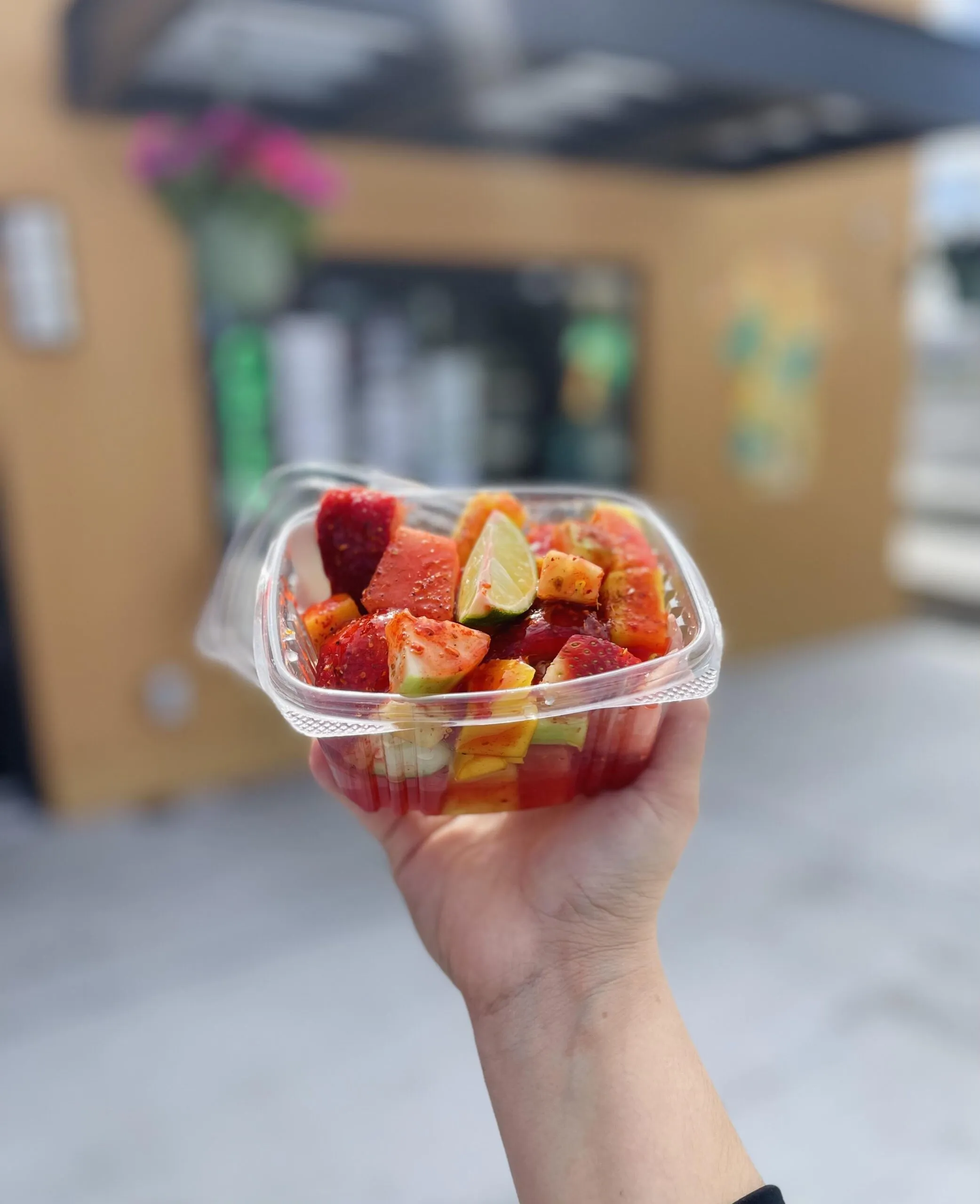 A hand holding a clear cup of fresh-cut fruit from the Tropical Express fruit bar in front of the shop