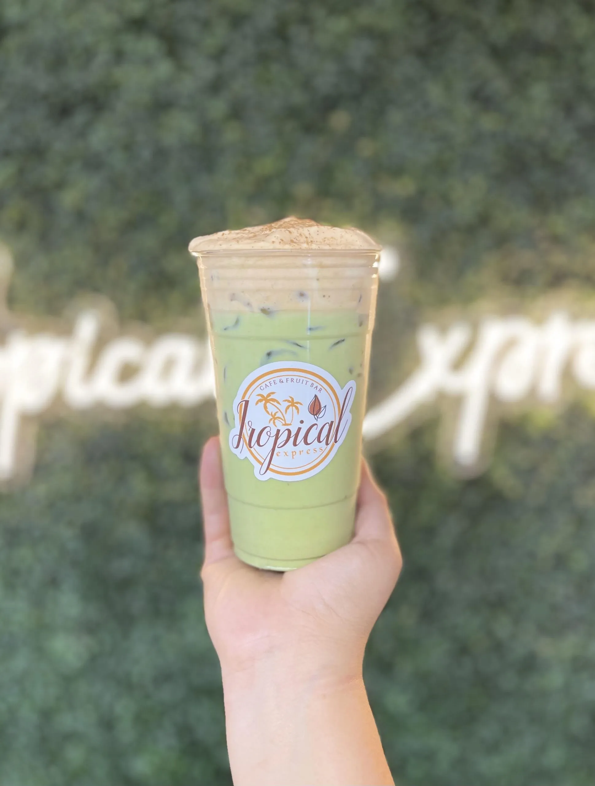 A hand holding an iced matcha latte in a Tropical Express cup in front of the brand’s neon script wall