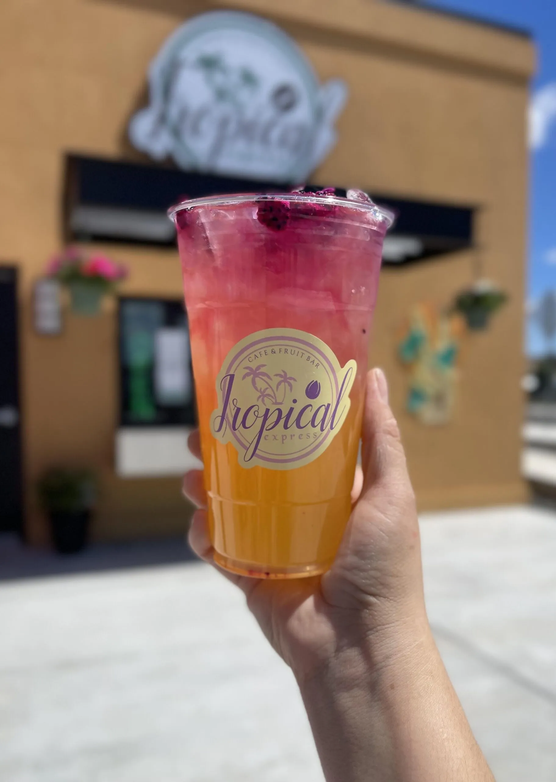A hand holding a pink-and-orange Tropical Express drink in front of the shop’s storefront on a sunny day